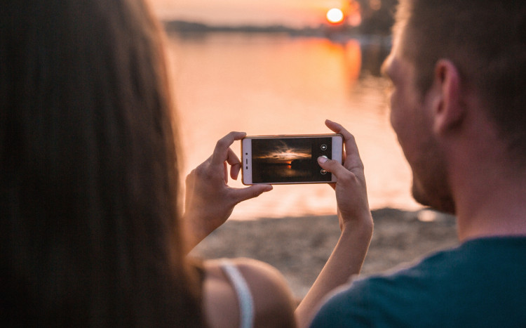 Kobieta i mężczyzna wykonujący zdjęcie smartfonem zachodu słońca nad wodą