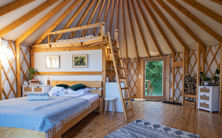 Wnętrze jurty, a w nim łóżko, drewniane wykończenie, mała kuchnia.