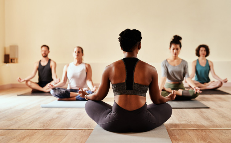 Jedna kobieta - trenerka - siedzi plecami i pokazuje pozostałym osobom w jaki sposób należy medytować