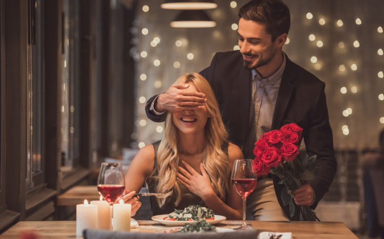 Mężczyzna zakrywa oczy siedzącej kobiecie i trzyma za nią bukiet róż