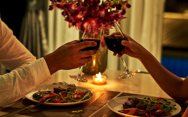 Dwie dłonie (męska i kobieca) trzymają kieliszek czerwonego wina w obecności kwiatów i świeczki