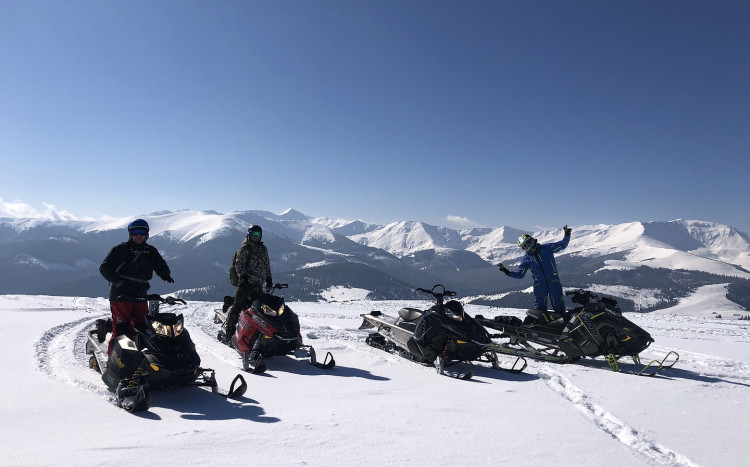 Cztery skutery śnieżne i trzy osoby pozujące do zdjęcia na tle gór pokrytych śniegiem