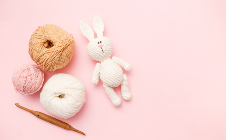 Wyszydełkowany królik w białym kolorze leżący na różowym tle, obok niego trzy włóczki: brązowa, biała i różowa oraz drewniane sz