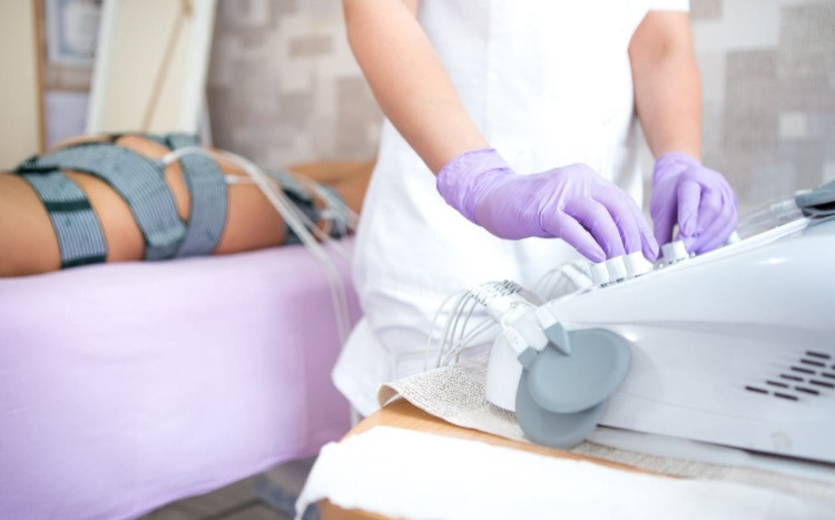 Kobieta w fioletowych rękawiczkach ustawia parametry urządzenia do masażu. Za nią kobieta z pasami na udach, pośladkach i biodra