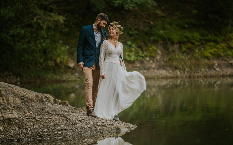 Kobieta w sukni ślubnej i mężczyzna w garniturze spacerujący nad brzegiem jeziora