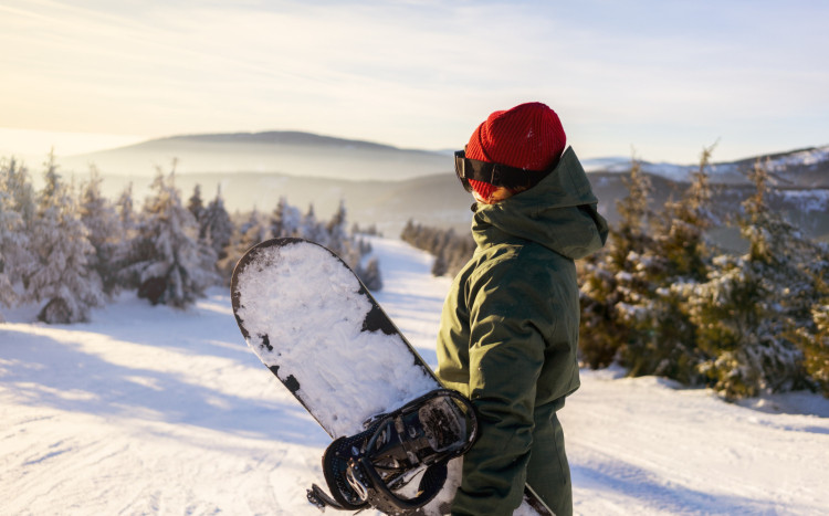 Osoba w czapce i goglach w górach pełnych śniegu trzymająca deskę do snowboardu