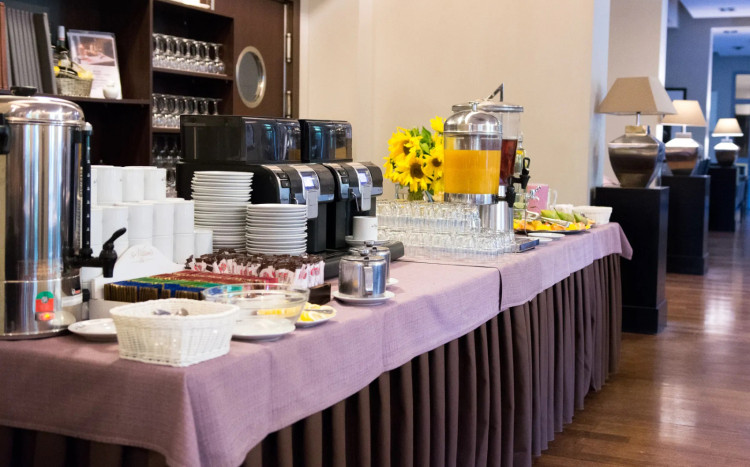 Stół, na którym znajdują się dwa ekspresy do kawy, talerzyki, szklanki, warnik do wody, herbaty i cukier