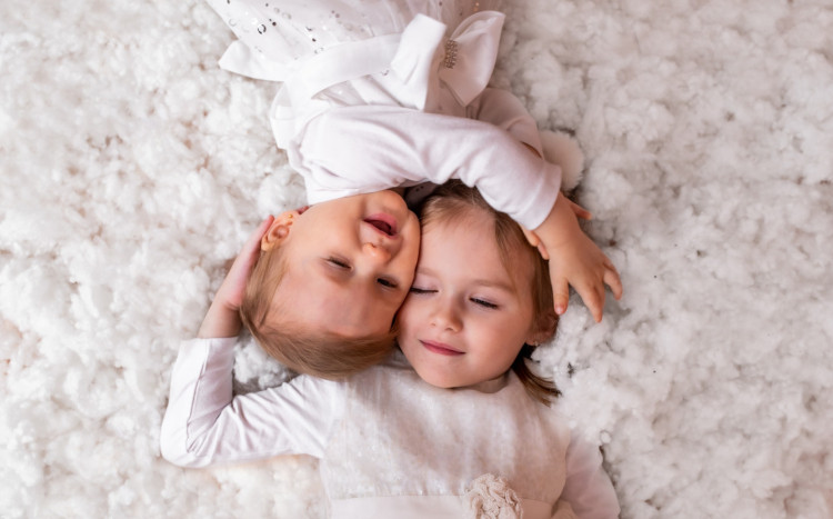 Dwie małe siostry przytulone do siebie głowami, ubrane w białe sukienki i leżące na białym puchu