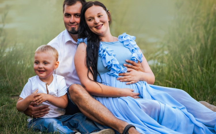 Szczęśliwa rodzina siedząca na trawie i pozująca do zdjęcia. Kobieta w niebieskiej sukience jest w ciąży