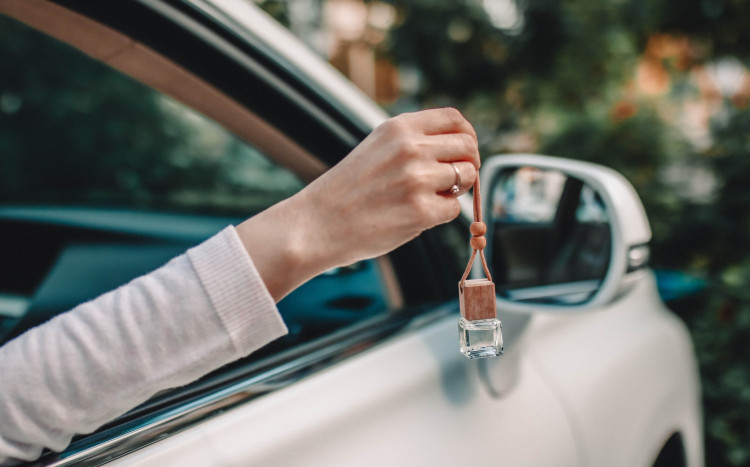 Damska dłoń z pierścionkiem trzymająca zapach do samochodu poza oknem na tle białego auta