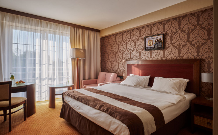 Pokój dwuosobowy urządzony w kolorach brązu z dwuosobowym łóżkiem, różowym fotelem, lampą, stołem i krzesłami