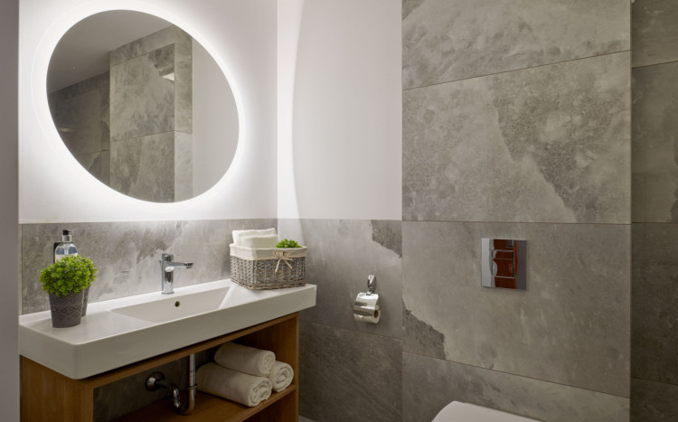 Łazienka, a w niej duże i okrągłe lustro z jasnym podświetleniem, umywalka, ręczniki oraz zielone rośliny